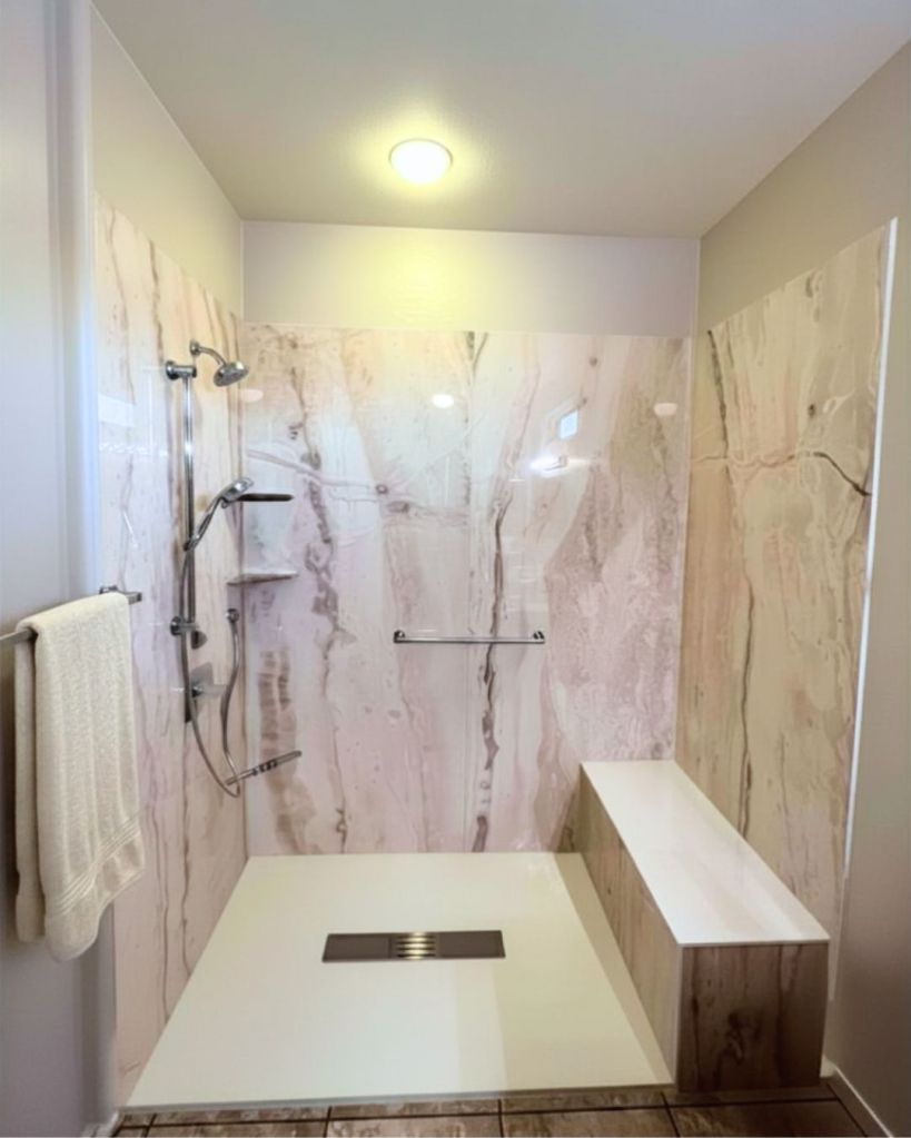 Accessible bathroom remodel by The Bath Authority with low-threshold walk-in shower, built-in bench seating, handheld showerhead, corner shelving, foot rest, and marble-look wall panels for aging-in-place safety.