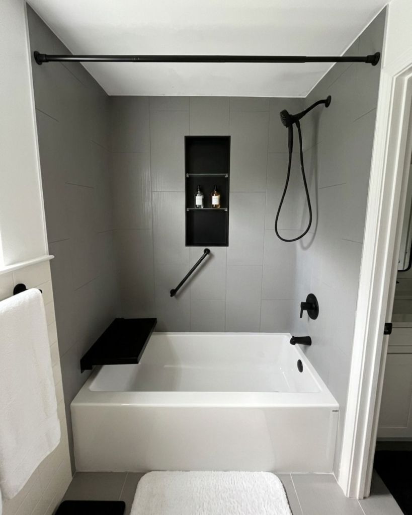 Modern bathtub replacement with gray wall surrounds, matte black shower fixtures, built-in bench seat, recessed storage niche, and grab bar installed by The Bath Authority.