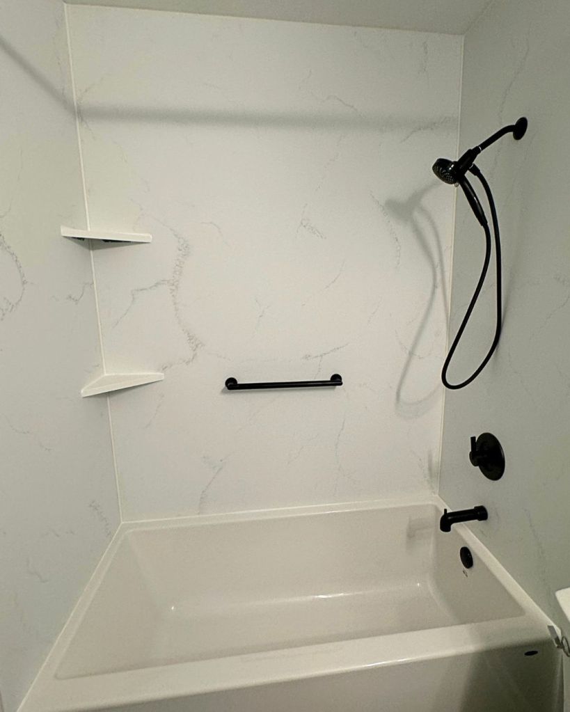 Bathtub and shower combination with white marble-look wall panels, matte black showerhead, corner storage shelves, and grab bar installed by The Bath Authority.