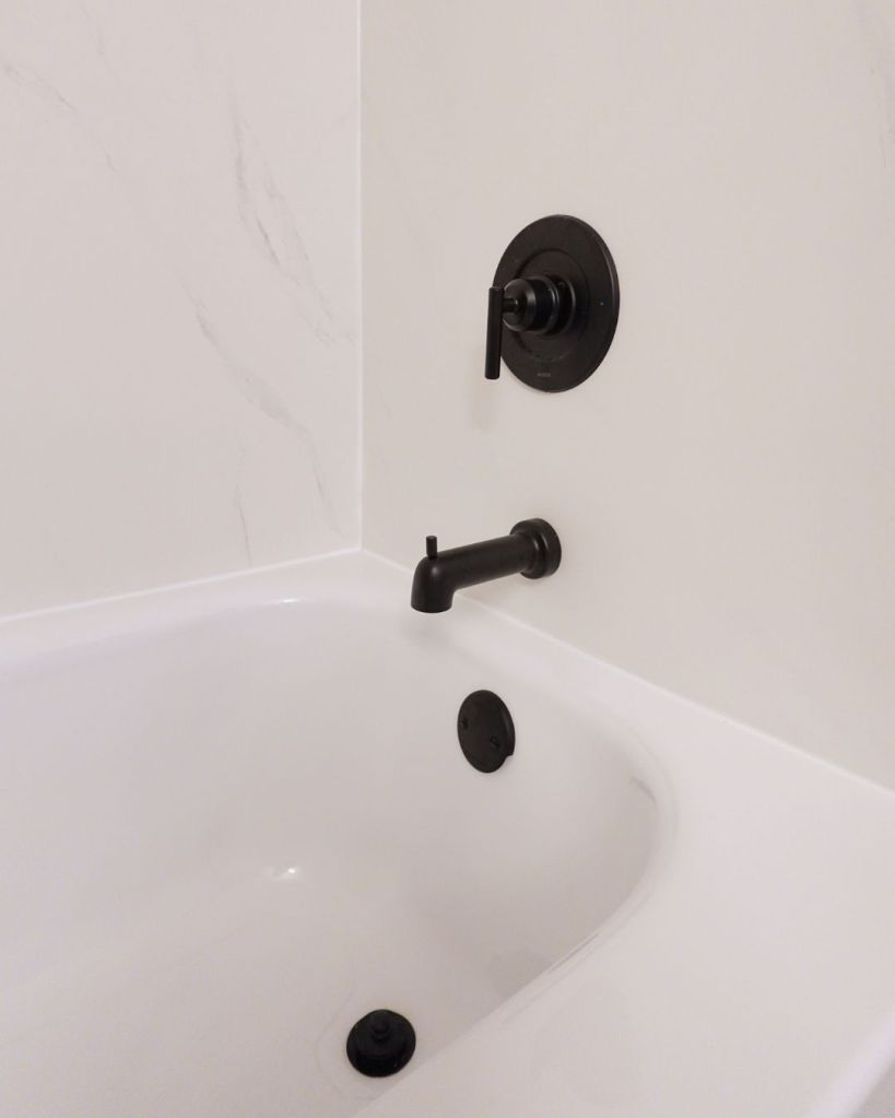 Close-up of a white bathtub with matte black faucet, drain hardware, and marble-look wall surround installed by The Bath Authority.