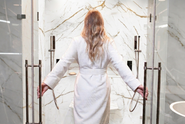 Homeowner stepping into a walk-in shower with white marble-look engineered stone walls and gold veining, designed by The Bath Authority.