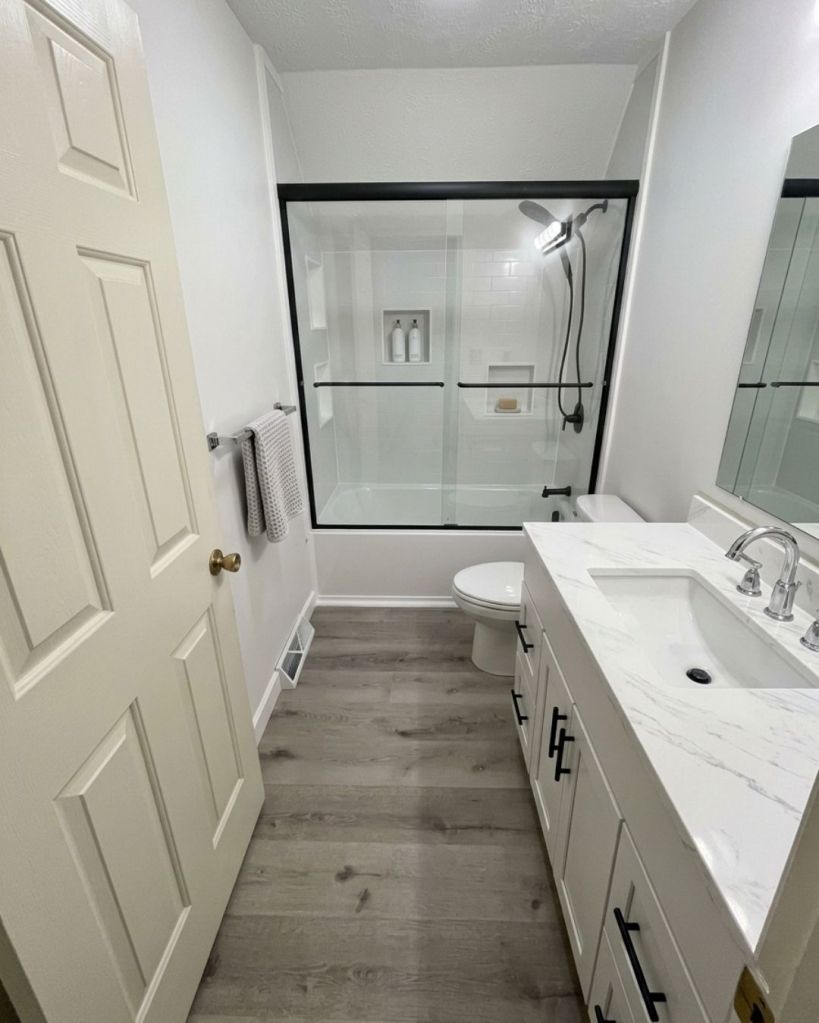 Contemporary full bathroom remodel completed by The Bath Authority featuring a custom white vanity, quartz countertop, luxury vinyl plank flooring, and bathtub-shower combination with black-framed glass doors.