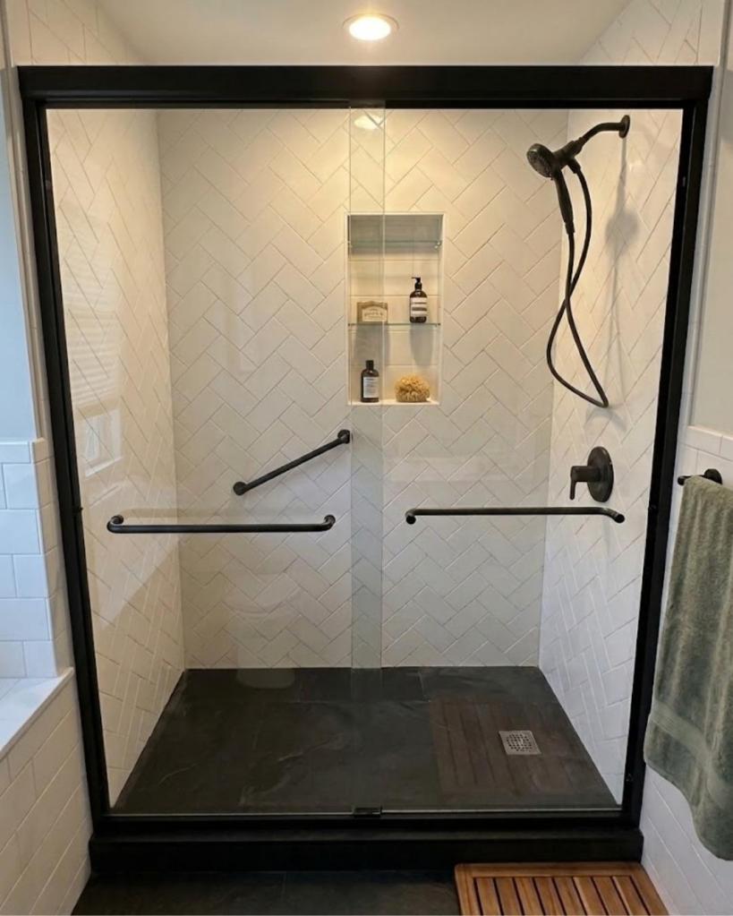 Custom walk-in shower with white herringbone walls, black-framed glass doors, grab bars, and built-in niche by The Bath Authority.