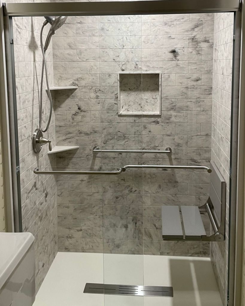 Accessible shower remodel completed by The Bath Authority with wide subway tile pattern gray wall panels, fold-down shower seat, grab bar, and recessed storage niche with corner shelves.