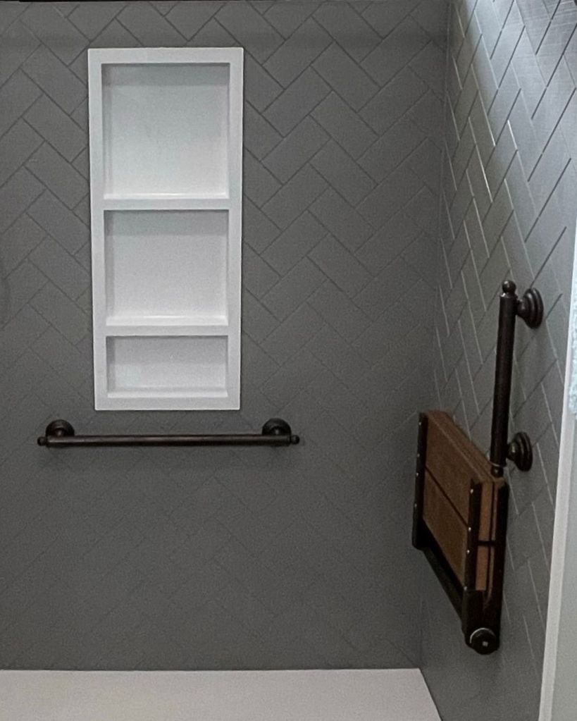 Walk-in shower completed by The Bath Authority featuring matte black hardware, a fold-down seat, large-format wall panels, and white built-in storage in a modern bathroom design.