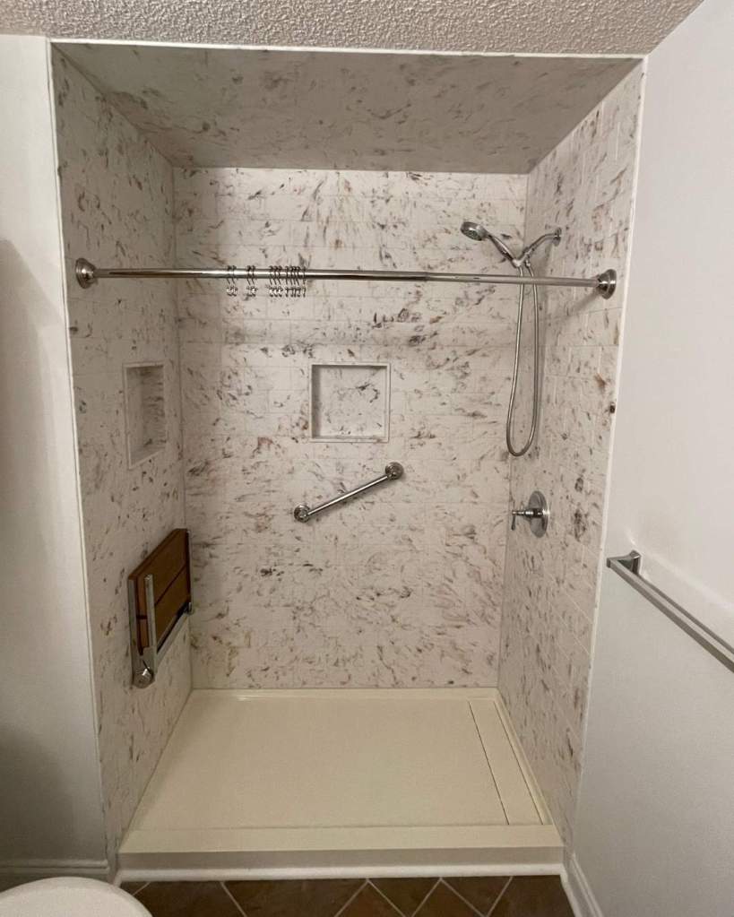 Accessible walk-in shower with speckled marble-look walls, chrome grab bars, bench seat, and handheld showerhead by The Bath Authority.