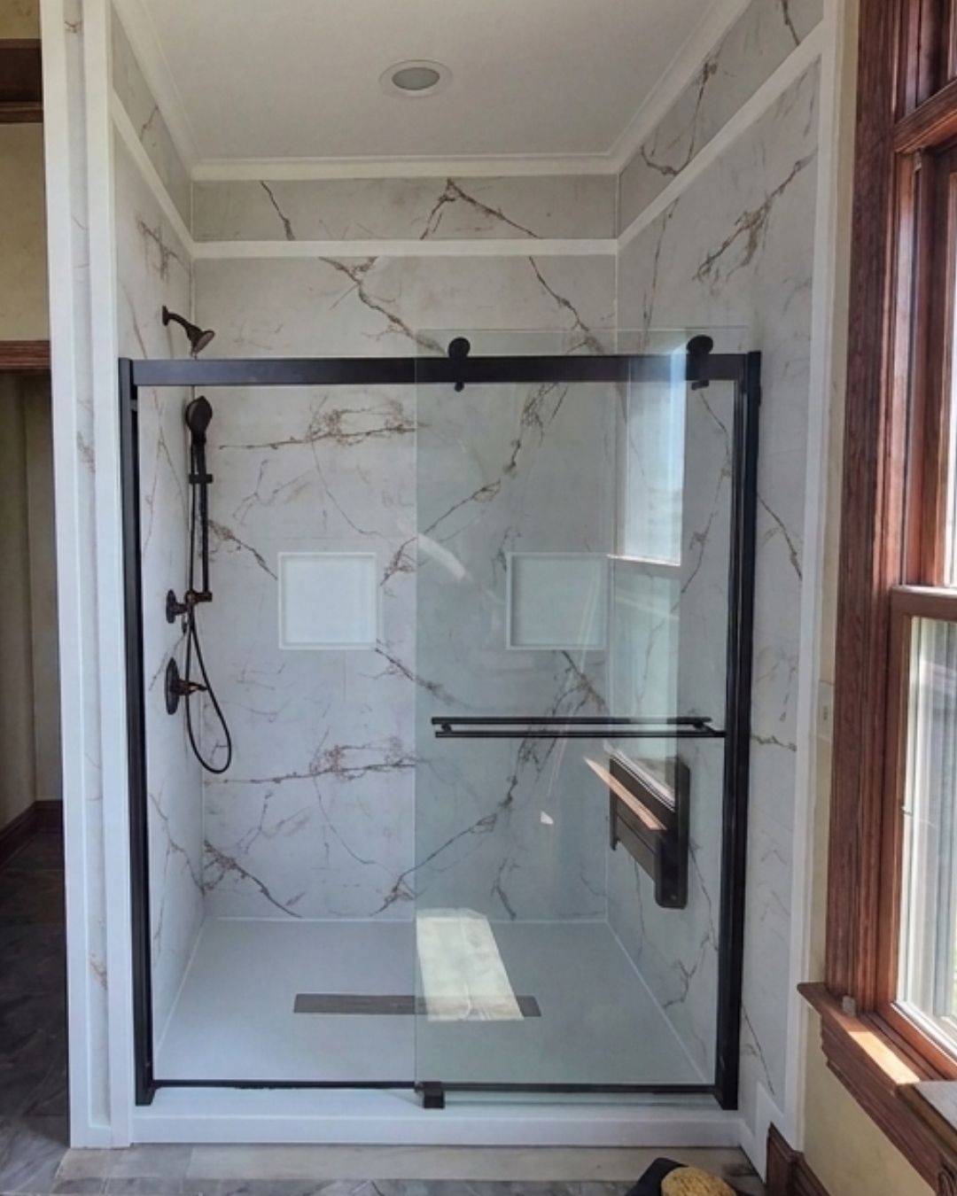 Luxury walk-in shower with marble-look walls, matte black barn-style glass door, and black shower fixtures by The Bath Authority.