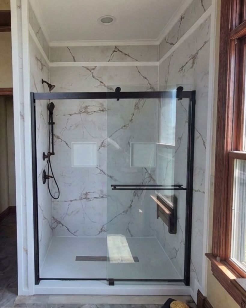 Luxury walk-in shower with marble-look walls, matte black barn-style glass door, and black shower fixtures by The Bath Authority.