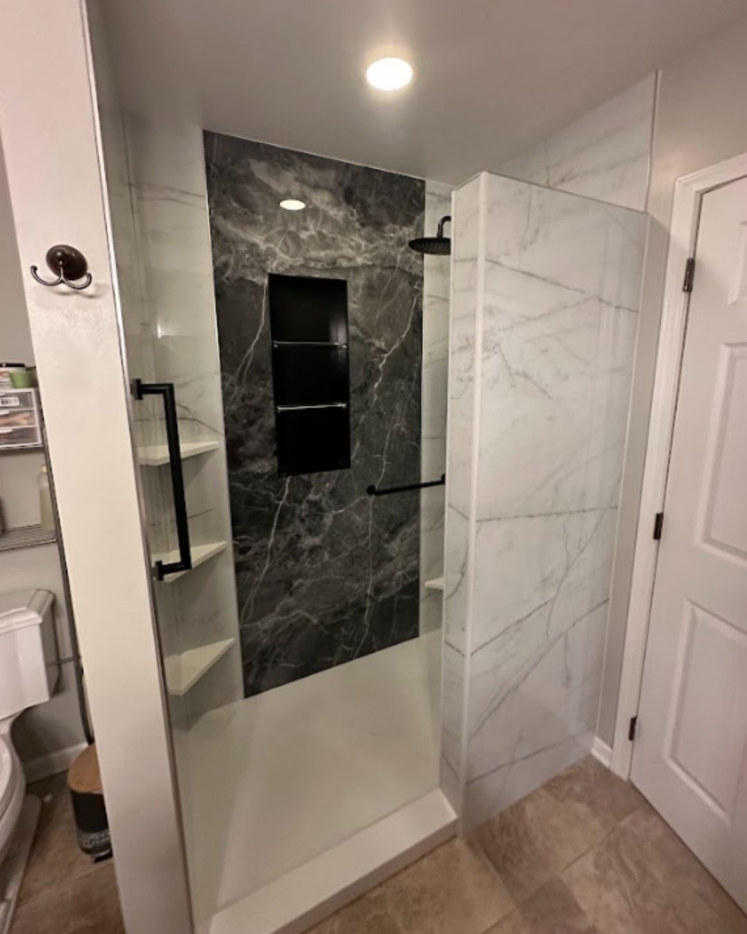 Open walk-in shower with white marble-look walls, dark marble accent wall, built-in shelves, and black fixtures by The Bath Authority.