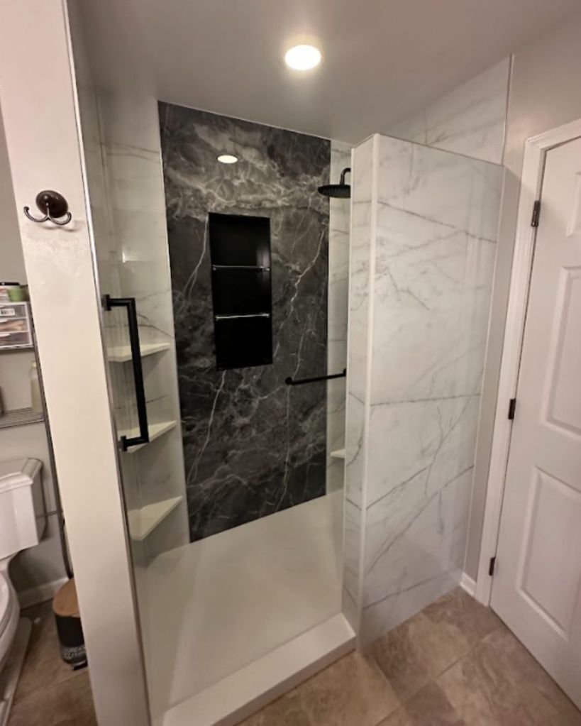 Open walk-in shower with white marble-look walls, dark marble accent wall, built-in shelves, and black fixtures by The Bath Authority.