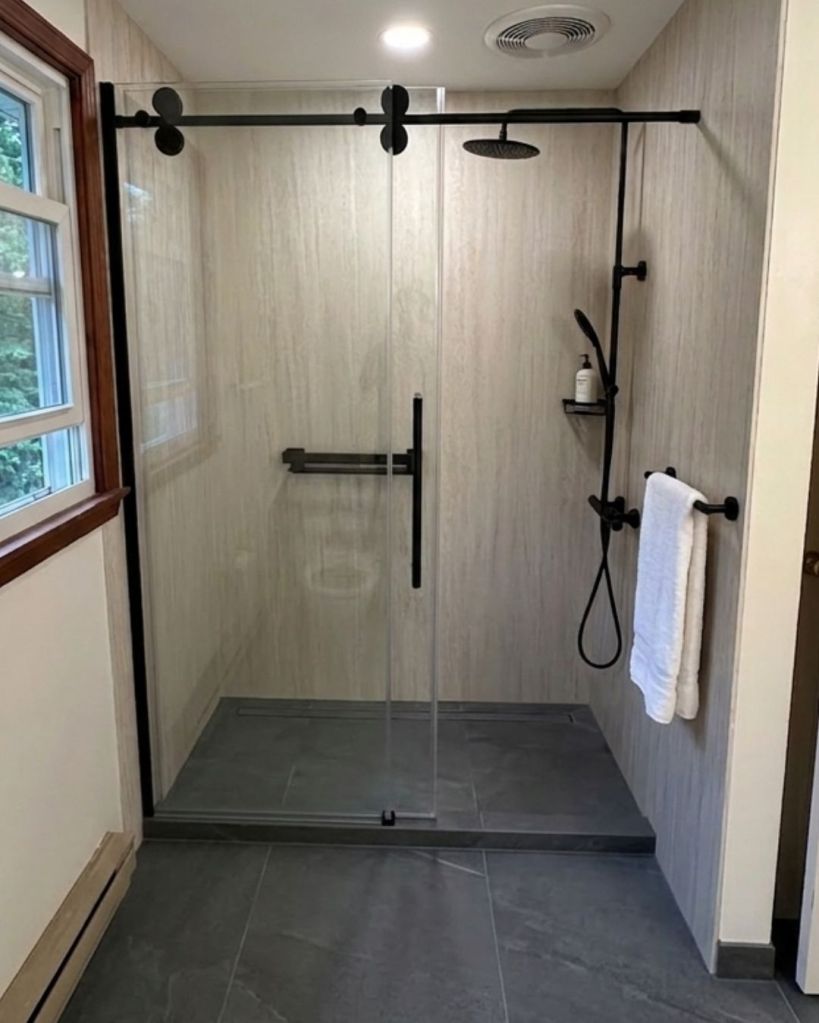 Spacious walk-in shower with stone-look wall panels, black sliding glass enclosure, rainfall showerhead, and handheld sprayer by The Bath Authority.