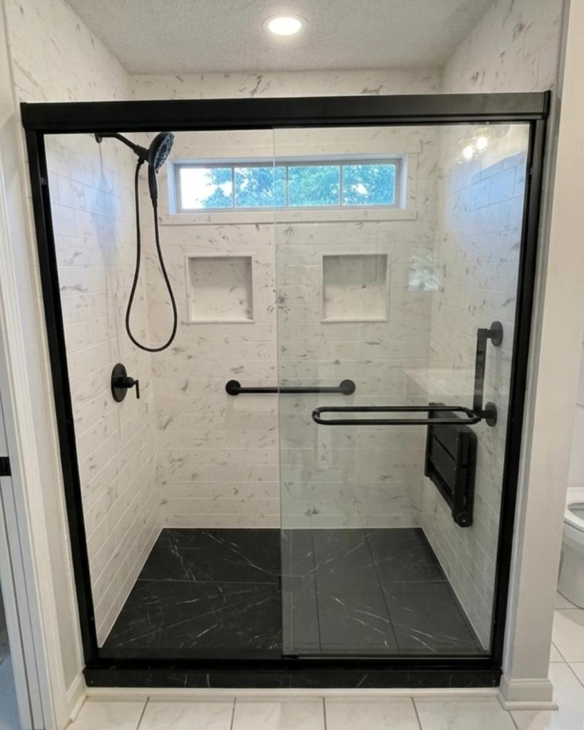 Walk-in shower with black-framed glass doors, marble tile-look walls, built-in niches, grab bars, and dark shower base by The Bath Authority.