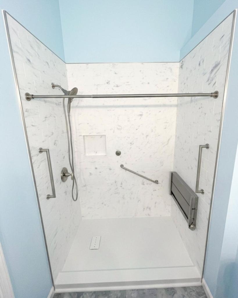 Accessible low-threshold walk-in shower with marble-look walls, grab bars, fold-down bench, and handheld showerhead by The Bath Authority.