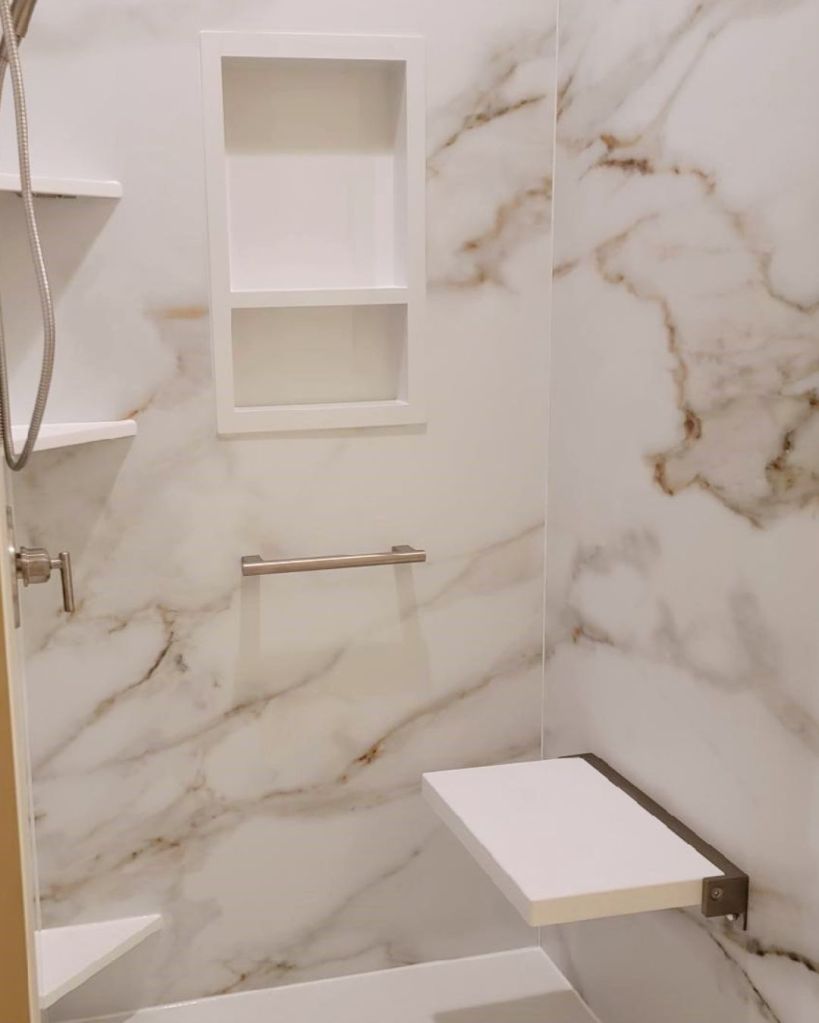 Walk-in shower interior with marble-look wall panels, recessed storage niche, corner shelves, and built-in shower seat by The Bath Authority.
