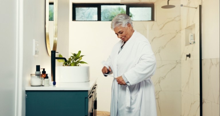 A senior woman stands smiling in her bathrobe in her bathroom.