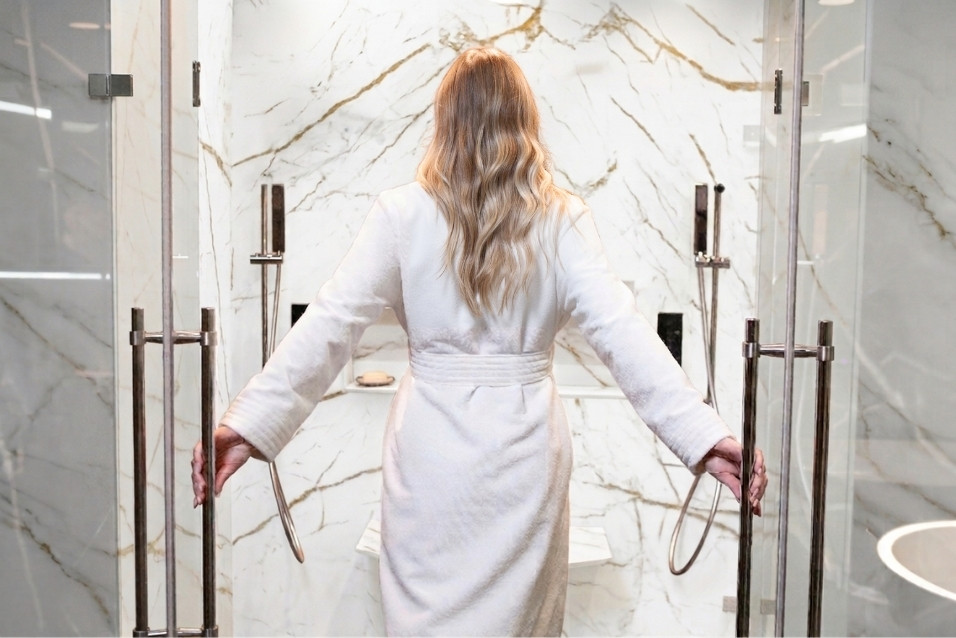 Homeowner stepping into a walk-in shower with white marble-look engineered stone walls and gold veining, designed by The Bath Authority.