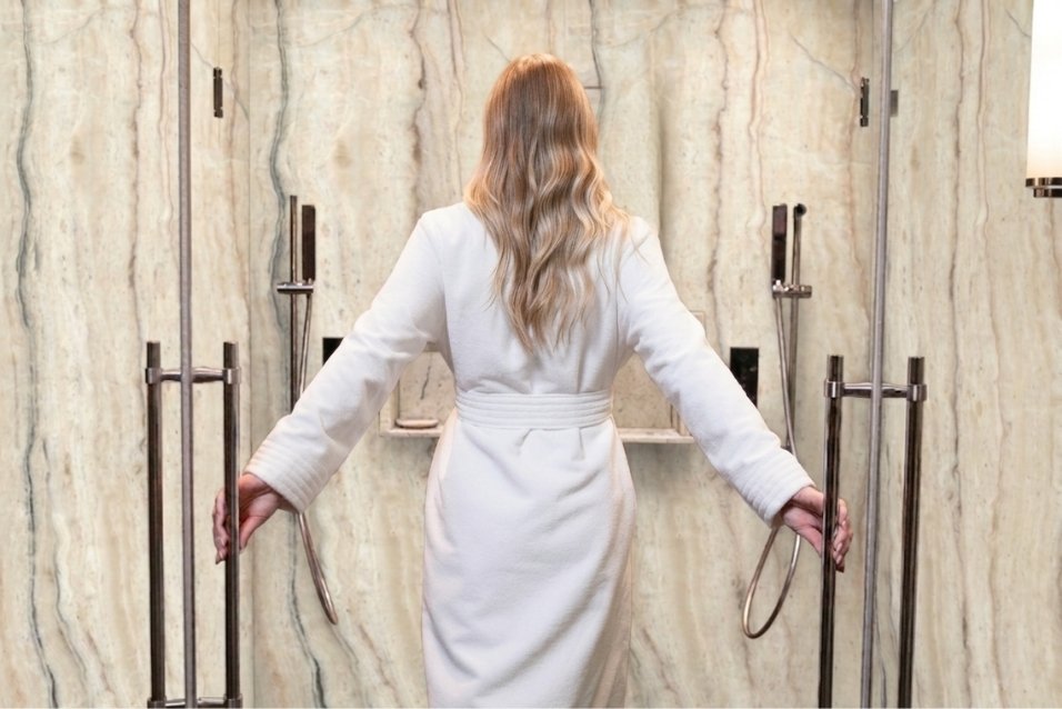 Homeowner entering a custom walk-in shower with warm cream-toned engineered stone wall panels installed by The Bath Authority.