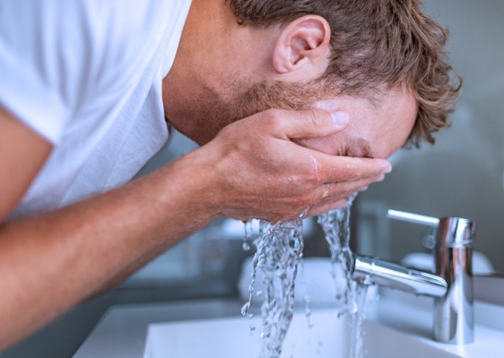 A man bends over a bathroom sink to splash water on his face.