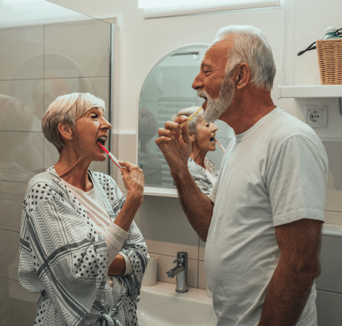 A senior man and woman stand in their bathroom brushing their teeth.