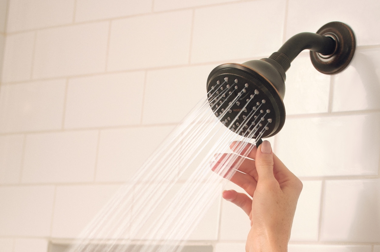 A hand adjusts a running showerhead on a white subway tile shower surround.